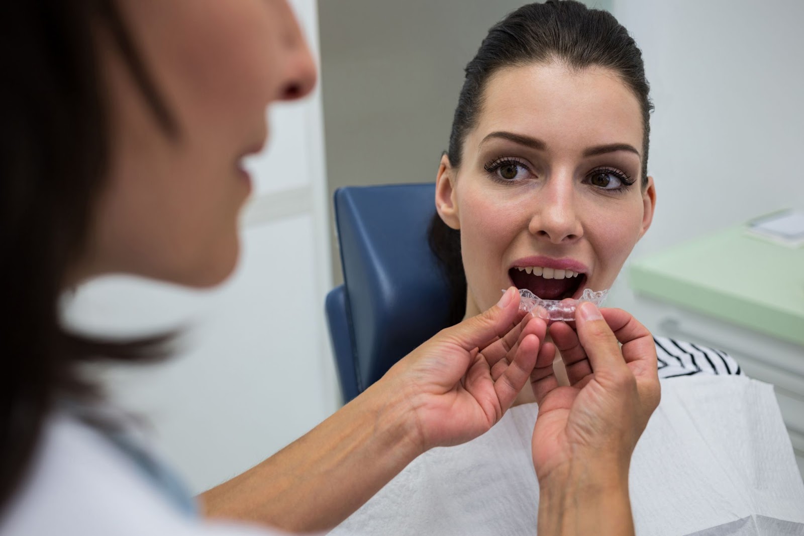 A woman sitting in a dental clinic chair opens her mouth as a dental professional holds a clear Invisalign aligner near her teeth, preparing to fit it.