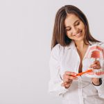 Woman demonstrating proper brushing technique using a dental model and toothbrush, smiling and promoting good oral hygiene.